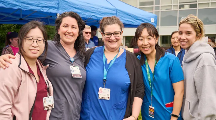 Five people, some in medical scrubs and wearing ID badges, stand together smiling outdoors at an event. Other people and a blue canopy tent are visible in the background.
