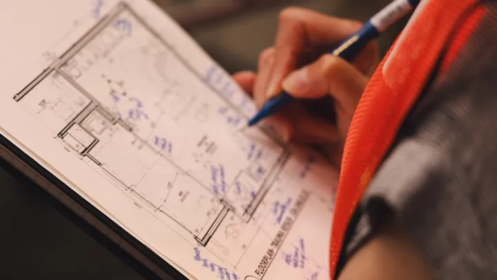 A close up of a clipboard showing architectural blueprints. A hand in the background is holding a blue pen and writing notes on the blueprints. 