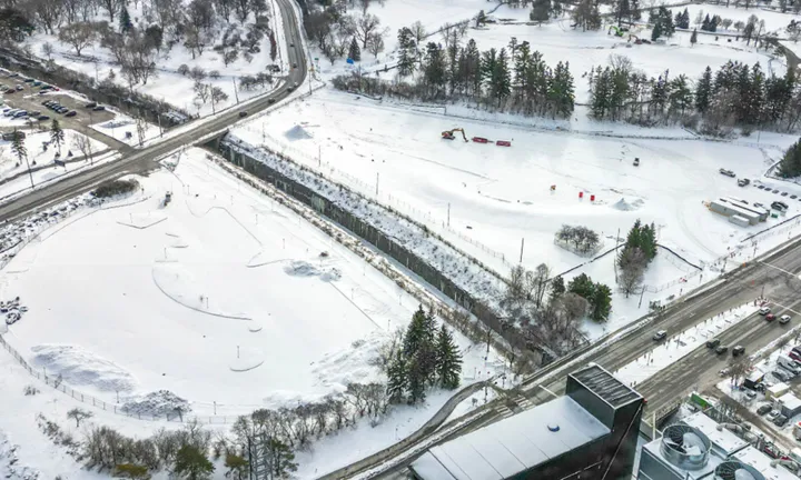 An aerial view in winter of the snow-covered construction site where the parking garage for The Ottawa Hospital’s new campus will be built. There are a few pieces of construction equipment, some trailers and a tractor on the site.