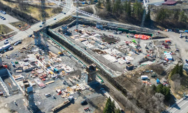 An aerial view in early spring of the construction site for the parking garage at The Ottawa Hospital’s new campus. Three large tower cranes are on the site. Large concrete structures like stairwells, elevator shafts and an entrance ramp for vehicles are partially built, extending vertically up from the ground level. 
