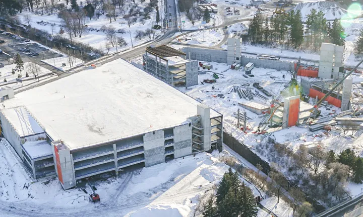 Vue aérienne en hiver du stationnement à étages partiellement construit sur le nouveau campus de L’Hôpital d’Ottawa. Une moitié de la structure est achevée et recouverte de neige, et les premières étapes de la construction ont commencé sur l’autre moitié.