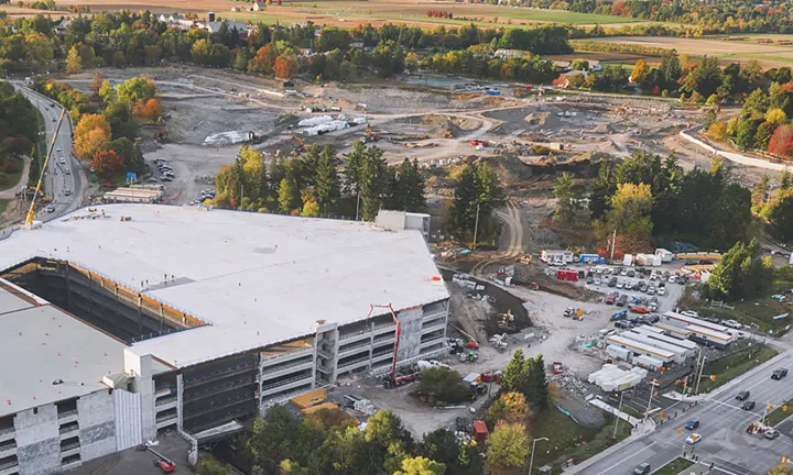 Vue aérienne du chantier de construction du nouveau campus de L’Hôpital d’Ottawa. Un stationnement à étages est en cours de construction en bas à gauche, et une vaste zone de terrain est en cours d’excavation en haut au centre. Le site est entouré d’arbres et de champs herbeux.