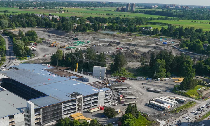 Vue aérienne du chantier de construction du nouveau campus de L’Hôpital d’Ottawa. Un stationnement à étages est en cours de construction en bas à gauche, et une vaste zone de terrain est en cours d’excavation en haut au centre. Le site est entouré d’arbres et de champs herbeux. 