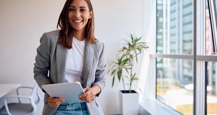 A person standing in a bright office space holds a tablet, with large windows and a potted plant in the background.