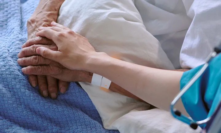 A medical professional gently holding the hand of a patient lying in a hospital bed.