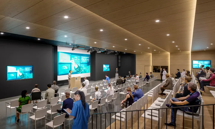 An artist rendering of the interior of an auditorium at a new hospital. A man is speaking at a podium; three large screens are on the wall behind him. There are multiple rows of chairs and various people seated throughout the large room.