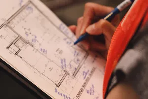 A close up of a clipboard showing architectural blueprints. A hand in the background is holding a blue pen and writing notes on the blueprints. 