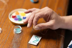 Gros plan de la main et des doigts d’une participante à l’atelier attrapant un bout de fil blanc. Il y a des piles de petites perles de différentes couleurs sur la table. On voit les premières étapes de ce qui deviendra un porte-clés en perles.