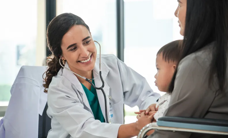 Une médecin utilise un stéthoscope pour examiner un jeune enfant assis sur les genoux d’une femme dans un cabinet médical.