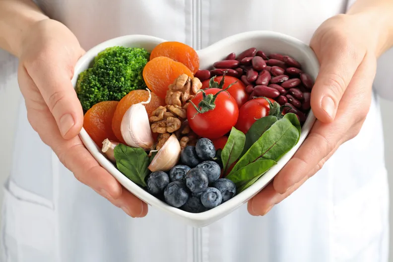 Une personne en blouse blanche tient un bol en forme de cœur rempli d’aliments sains, notamment du brocoli, des abricots secs, des noix, de l’ail, des tomates cerises, des haricots rouges, des épinards et des bleuets.