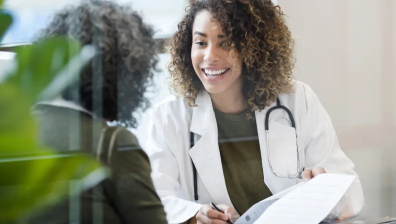 Une médecin souriante aux cheveux bouclés qui porte un stéthoscope autour du cou s’entretient avec une patiente tout en tenant un document au cours d’une consultation médicale qui semble positive et conviviale.