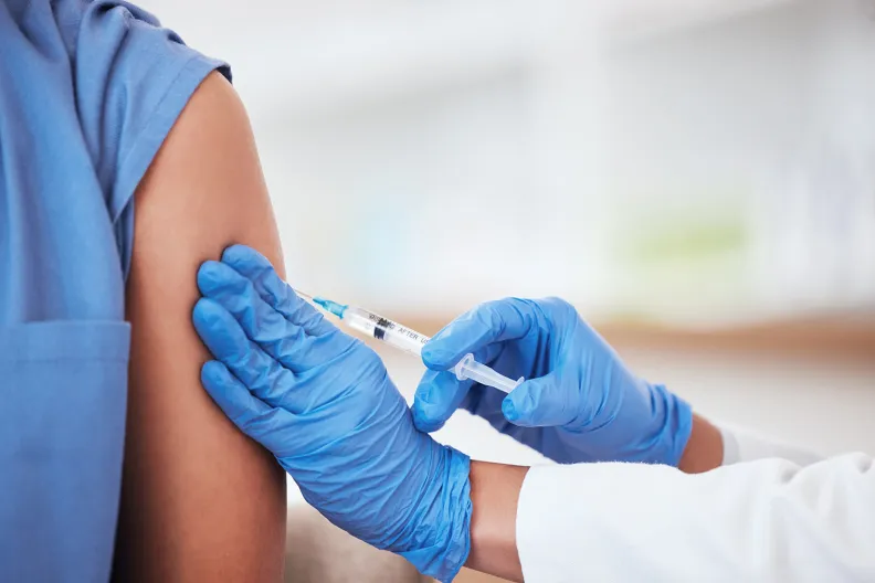 Un professionnel de santé portant des gants bleus administre un vaccin dans le bras d’un patient. Le patient porte une manche bleue. L’arrière-plan est flou.