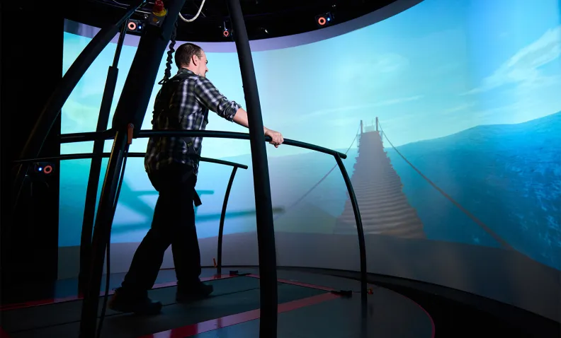 Une personne utilise un tapis roulant de réalité virtuelle. Dans le cadre de cette expérience immersive, elle marche vers une image de pont suspendu projetée sur un écran incurvé.
