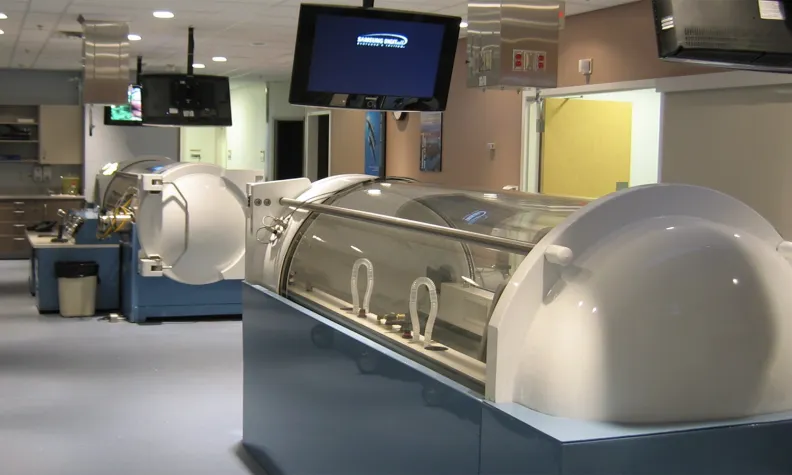 A modern medical room with large hyperbaric oxygen therapy chambers, overhead monitors, and various medical equipment, featuring clean, bright lighting and a tidy environment.
