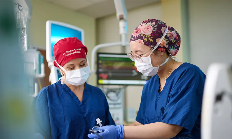 Deux professionnelles de la santé vêtues de tenues de chirurgie, de masques et de gants effectuent une intervention sur un patient dans une salle d’un hôpital. Il y a des écrans et du matériel médical en arrière-plan.