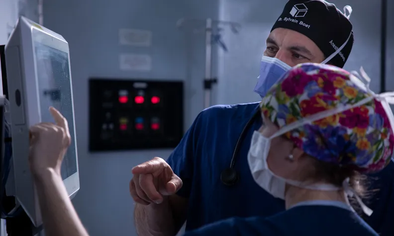 Two medical professionals in scrubs and masks look at and point to a monitor in a hospital setting, appearing to discuss or review information on the screen.