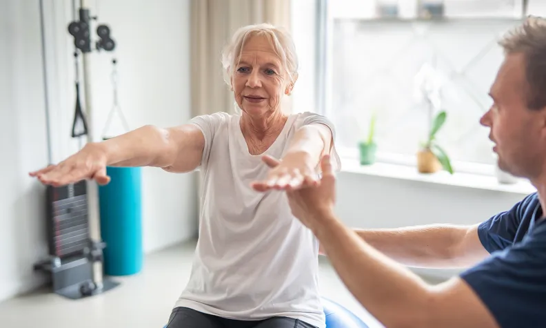An older woman sits on an exercise ball with arms extended forward, performing a balance exercise. A physical therapist kneels beside her, offering support and guidance in a bright, modern room.