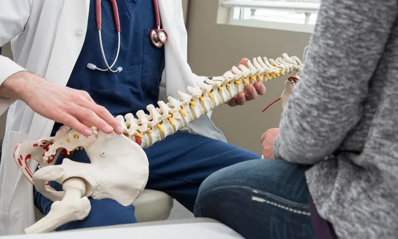 A doctor in a white coat holds and explains a model of the human spine to a patient seated nearby in a medical office.