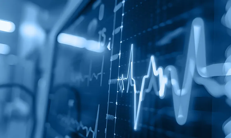 A close-up of a medical heart rate monitor display shows a glowing ECG waveform and vital signs in a hospital setting, with a blue-toned, blurred background.