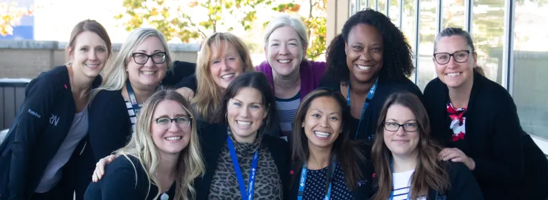 A smiling group shot of ten clinical workers, six in the back and four in the front, huddled together in a sunny outdoor setting, near a wall of windows. All are in dark blue medical scrubs