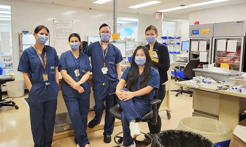 Group of TOH Pharmacy Environmental Stewardship committee members standing and seated together in a hospital pharmacy workspace.