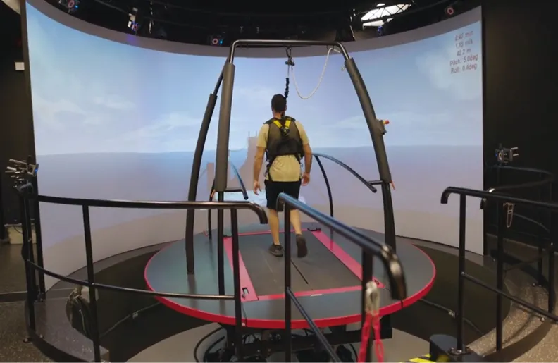 Une personne portant un harnais de sécurité marche sur un grand tapis roulant circulaire à l’intérieur d’un environnement immersif de réalité virtuelle, entourée d’écrans courbés affichant une scène extérieure.