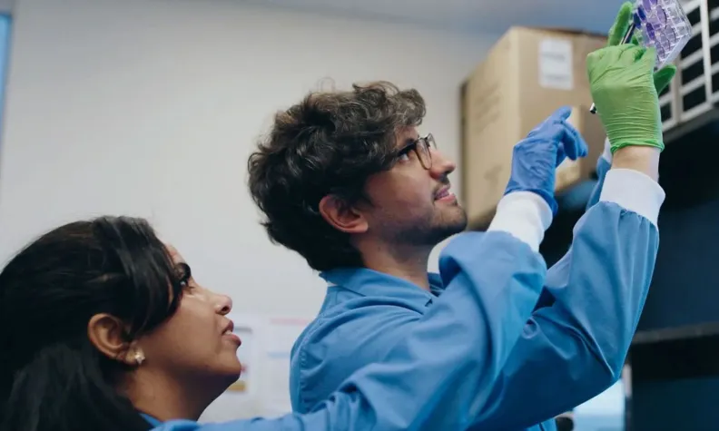 Two researchers in lab coats work together in a laboratory. One person holds up a clear multi‑well plate with gloved hands, examining it closely. The other person stands nearby, observing. 