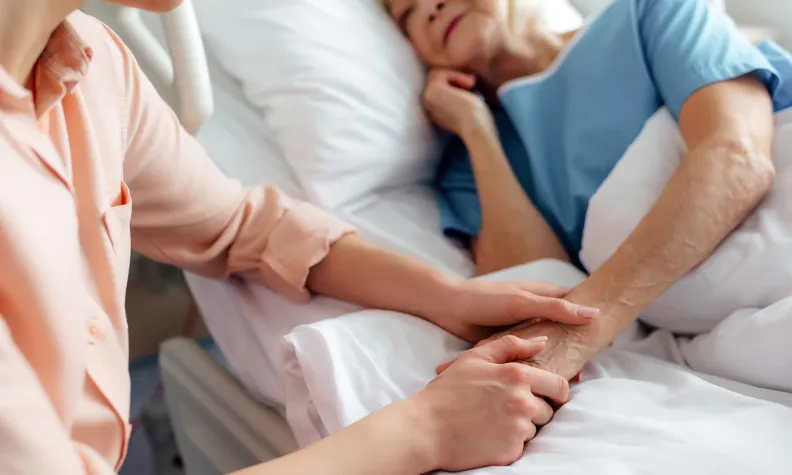 A person sits beside a hospital bed, gently holding the hand of an elderly patient who is lying under white blankets, wearing a blue hospital gown and appearing comforted by the support.