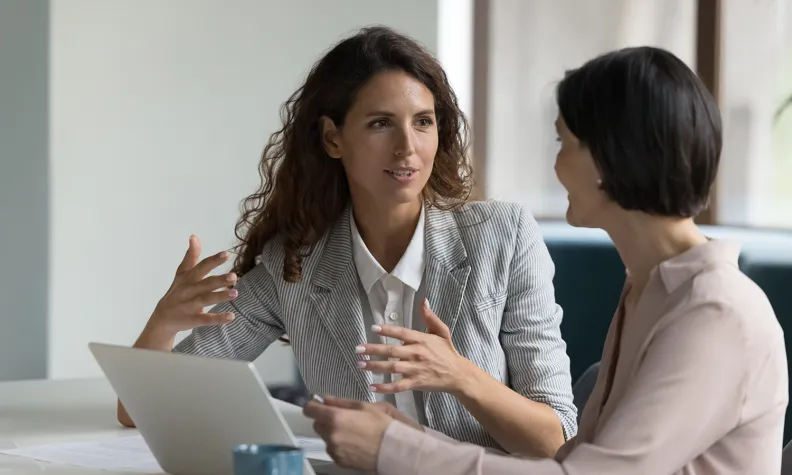 Two people sit at a table discussing something; one gestures with their hands while the other listens, holding a tablet. A laptop, documents, and a blue mug are on the table in a modern office setting.