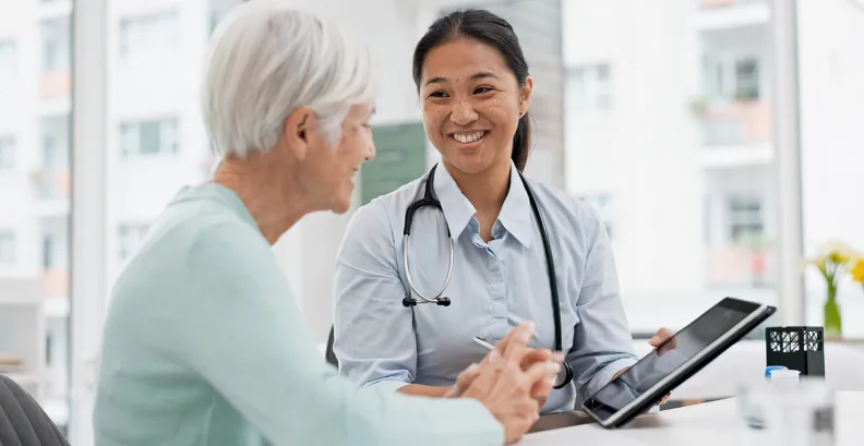 A doctor holding a tablet smiles while talking with an older patient in a medical office. They are seated at a desk, suggesting a friendly and informative consultation.