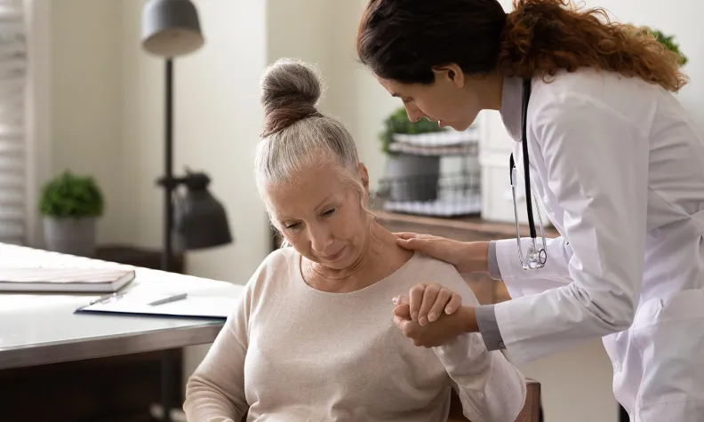 A doctor comforts an elderly person by holding their hand and touching her shoulder. The person looks sad or concerned as they sit in a chair in a well-lit room.