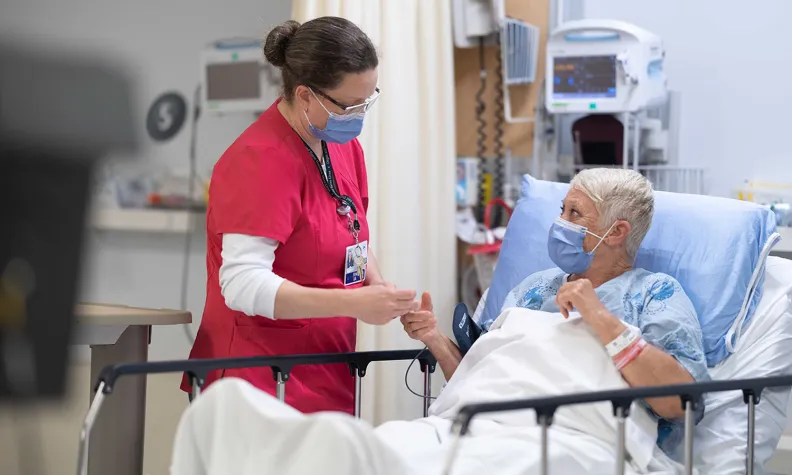 A nurse in red scrubs wearing a mask stands next to a hospital bed, holding the hand of an elderly patient who is also masked and lying in bed with medical equipment in the background.