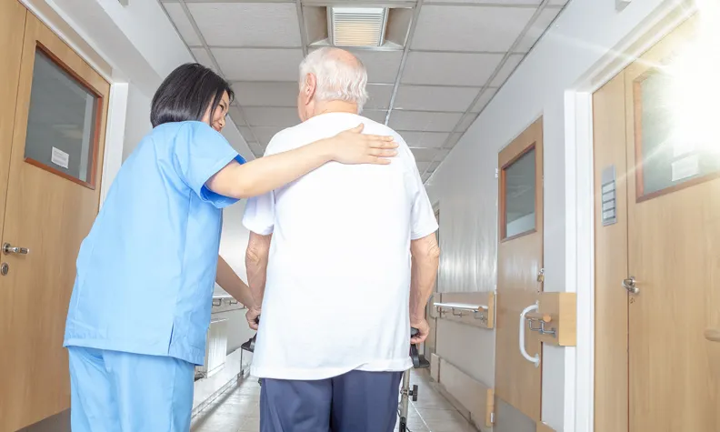 A nurse in blue scrubs supports and guides an elderly person using a walker as they walk down a bright hospital hallway together.