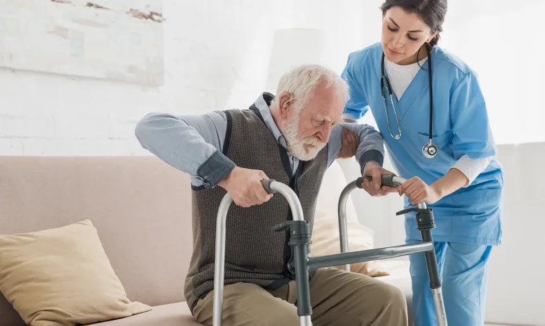 A nurse assists an elderly person using a walker as he stands up from a couch. The nurse is wearing blue scrubs and a stethoscope, offering support and care in a bright room.