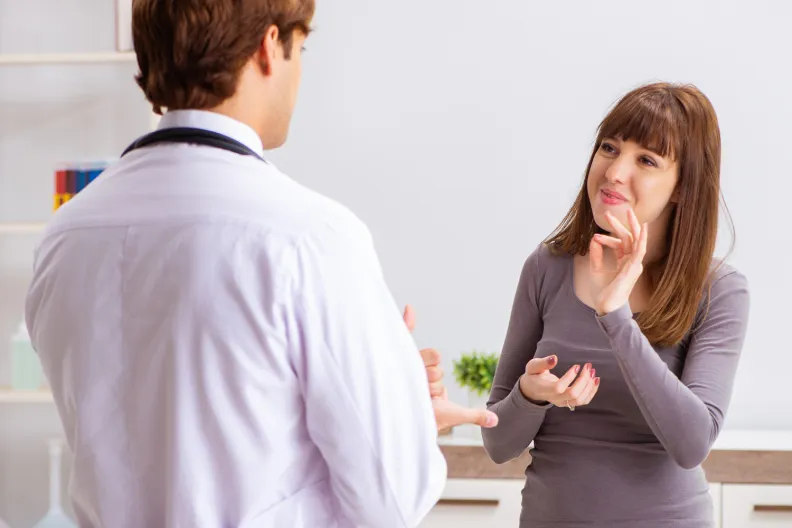 Une personne utilise la langue des signes pour communiquer avec un médecin dans un bureau bien éclairé. Le médecin fait face à cette personne et lui consacre toute son attention tandis que celle-ci fait des gestes et semble engagée dans une conversation.