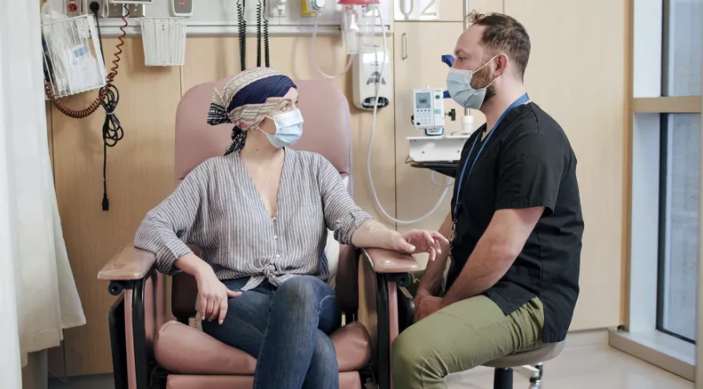 Une personne portant un foulard sur sa tête et un masque est assise sur une chaise d’hôpital. Elle tient la main d’un professionnel de la santé portant un uniforme médical et un masque.