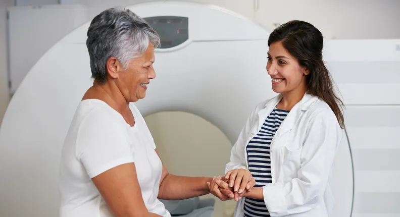 A healthcare professional in a white lab coat holds the hand of an older patient seated beside an MRI scanner, offering reassurance in a clinical imaging room.