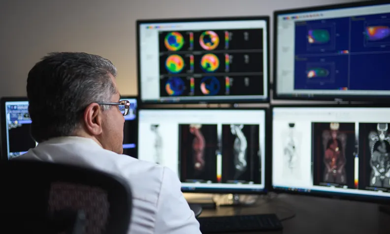 A medical professional in a white coat reviews PET scan and imaging results on multiple computer monitors.