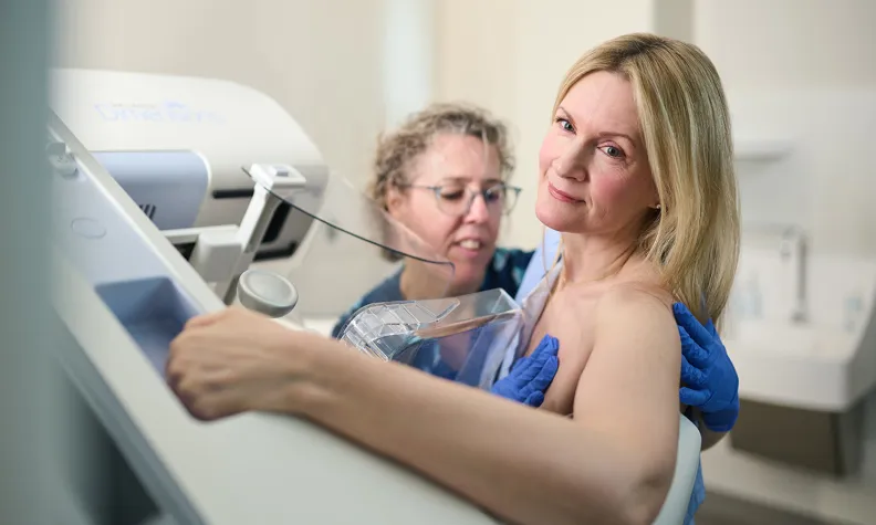 A healthcare professional assists a person during a mammogram screening, positioning their breast on the machine. The person looks slightly over their shoulder while the procedure is being prepared in a medical setting.