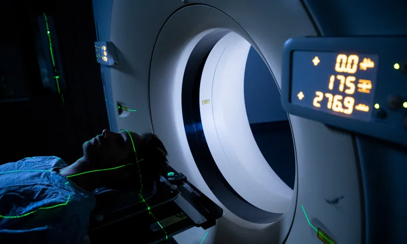A person lies on a table entering a CT scanner, with green laser guide lines on their head and display panels showing numbers on the machine.