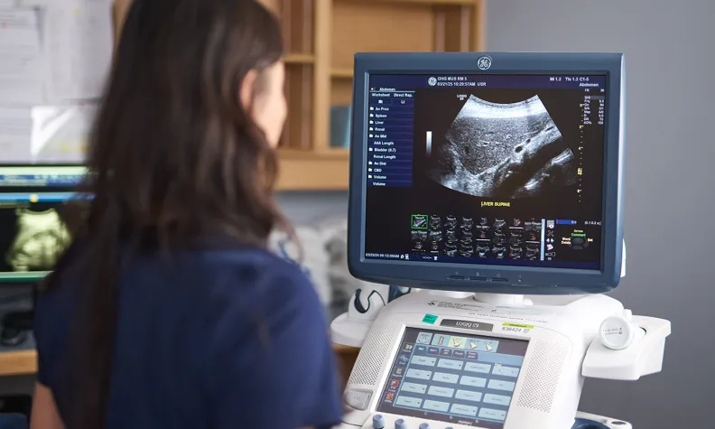 A person with long dark hair views an ultrasound image displayed on a monitor in a medical setting, with various control buttons and screens visible on the ultrasound machine.