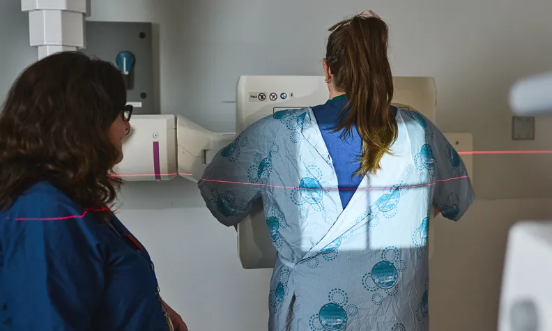 A person in a hospital gown stands facing a medical imaging machine, with a technician beside them. A red alignment laser is visible across their back, indicating a diagnostic procedure.