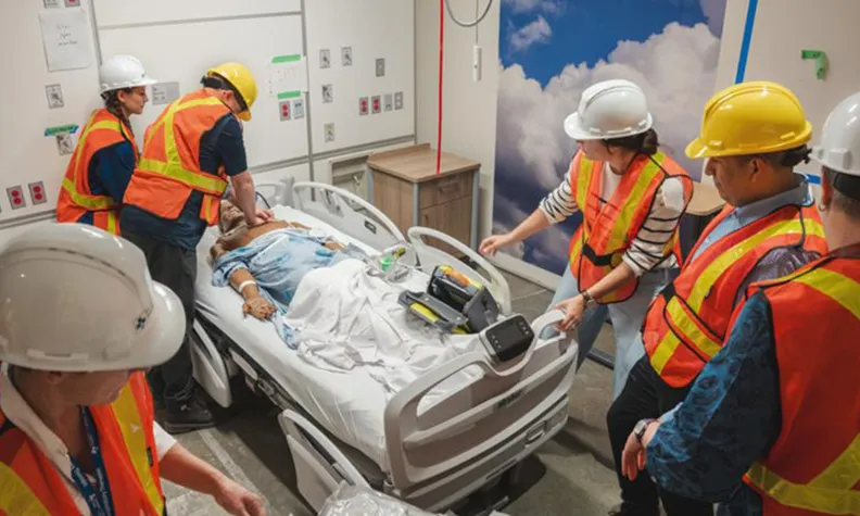 A high angle view of hospital staff conducting a simulation of a medical emergency. The staff are wearing orange safety vests and safety helmets and are surrounding a stretcher inside a temporary patient room. Two staff are performing simulated chest compressions on an artificial patient.