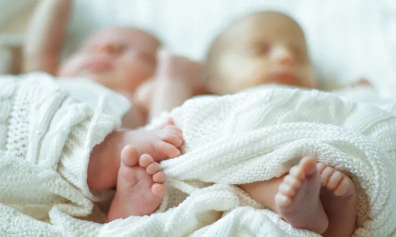 Two newborn babies lie side by side, wrapped in soft white blankets. Their faces are slightly out of focus, with their tiny feet prominently shown in the foreground.