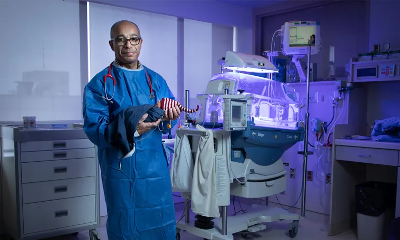 A doctor in blue scrubs and a stethoscope stands in a hospital room holding a small blanket. Behind him, there is a neonatal intensive care incubator with medical equipment.