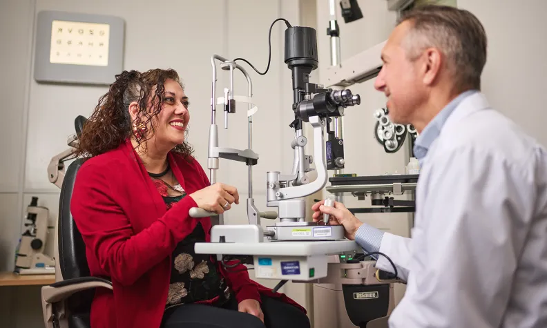 A person wearing a red jacket smiles while talking to an eye doctor during an eye exam. They are seated in an exam room with ophthalmology equipment and an eye chart visible in the background.
