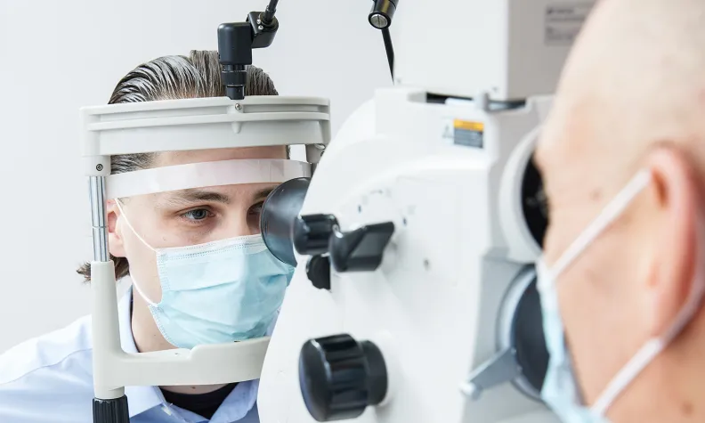 A patient wearing a mask undergoes an eye examination with specialized equipment while an eye care professional, also masked, operates the machine in a clinical setting.