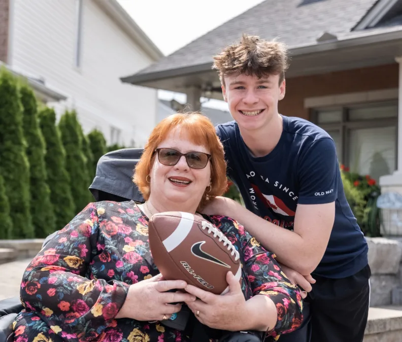 Une femme assise dans un fauteuil roulant tient un ballon de football, tandis qu’un jeune homme se tient près d’elle, un bras posé sur ses épaules. Ils sont à l’extérieur, dans un quartier résidentiel, devant une maison bordée de verdure.