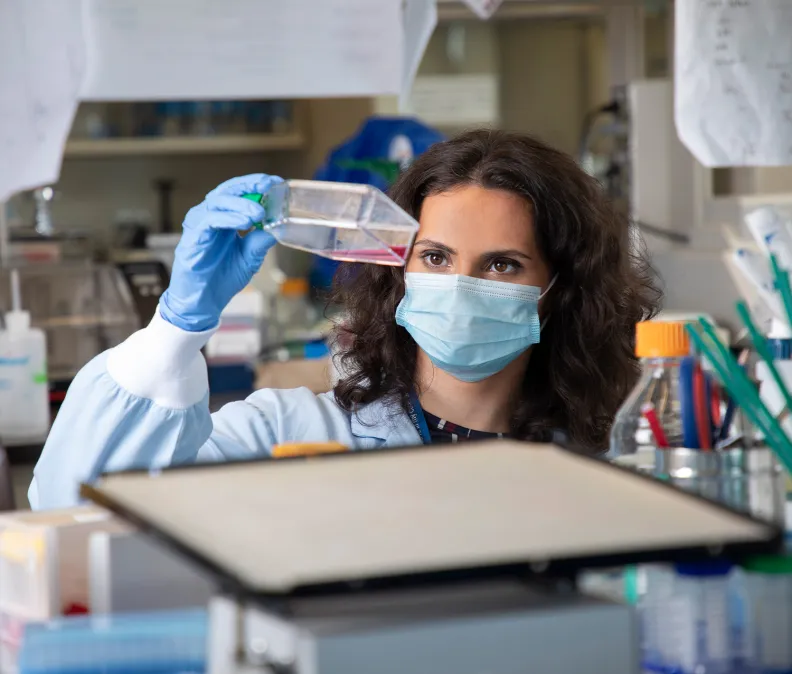 Dans un laboratoire, une chercheuse portant une blouse et des gants bleus tient un contenant de milieu rose et l’examine attentivement. Des équipements scientifiques, des bouteilles et divers outils sont visibles dans l’espace de travail.