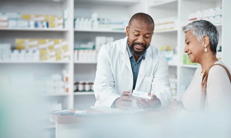 A pharmacist in a white coat explains medication instructions to a smiling older person at a pharmacy counter, with shelves of medicine in the background.